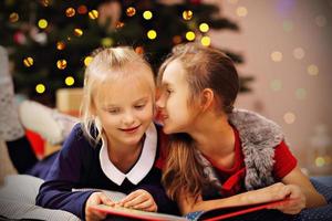 Two cute little sisters reading story book together under Christmas tree photo