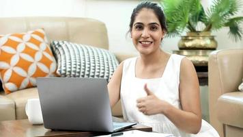 femme indienne souriante travaillant sur un ordinateur portable et montrant le geste du pouce levé.