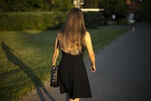 Girl in black dress walks through park. Girl with long hair in summer on street. Black dress and handbag. photo