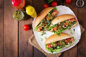 Hot dog with jalapeno peppers, tomato, cucumber and lettuce on wooden background photo