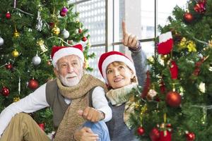 pareja caucásica mayor celebrando la navidad juntos en felicidad y emoción en casa con sombrero de santa y árbol de navidad foto