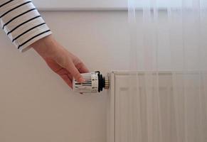 Woman regulates temperature at home with the heating thermostat to save energy, close-up with hand. Temperature control valve on radiator. White heater on wall with curtain in modern apartment. photo