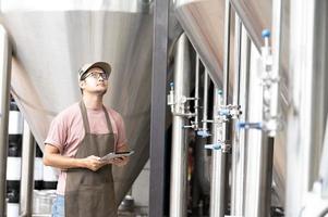 joven trabajador asiático que inspecciona la calidad de la cervecería con un vaso de cerveza artesanal que evalúa la apariencia visual después de la preparación mientras trabaja en una cervecería artesanal de procesamiento. foto