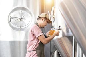 Young Asian worker inspecting brewery quality with a glass of craft beer evaluating visual appearance after preparation while working in a processing craft brewery. photo