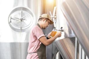 joven trabajador asiático que inspecciona la calidad de la cervecería con un vaso de cerveza artesanal que evalúa la apariencia visual después de la preparación mientras trabaja en una cervecería artesanal de procesamiento. foto