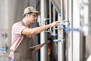 Young Asian worker inspecting brewery quality with a glass of craft beer evaluating visual appearance after preparation while working in a processing craft brewery. photo