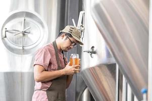 joven trabajador asiático que inspecciona la calidad de la cervecería con un vaso de cerveza artesanal que evalúa la apariencia visual después de la preparación mientras trabaja en una cervecería artesanal de procesamiento. foto