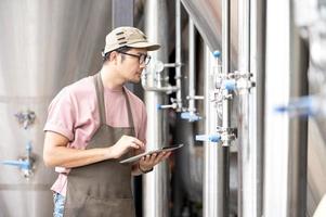 Young Asian worker inspecting brewery quality with a glass of craft beer evaluating visual appearance after preparation while working in a processing craft brewery. photo