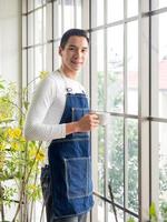 Gardener adult young Asian man one person handsome standing think look and Smiling  relaxing happy drinking a cup of coffee in the room cafe with glass window and green trees in spring morning time. photo
