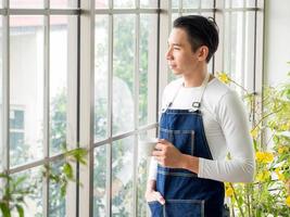 Gardener adult young Asian man one person handsome standing think look and Smiling  relaxing happy drinking a cup of coffee in the room cafe with glass window and green trees in spring morning time. photo