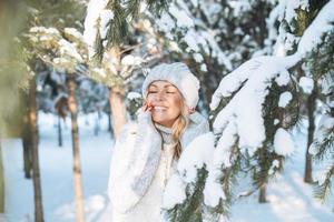 mujer rubia sonriente en ropa de invierno manos cerca de la cara en el bosque de invierno cubierto de nieve foto