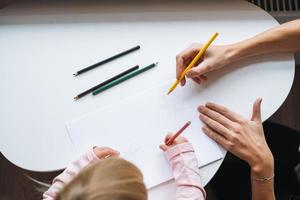 Little girl toddler with her mother drawing with colored pencils on table in children's room at home, view from top photo