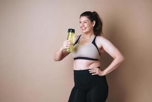 Self loving young woman plus size in sport with bottle of water on beige background, body love photo