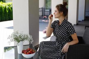 hermosa mujer con un hermoso vestido con un patrón de lunares sentado en un patio y comiendo fresa foto