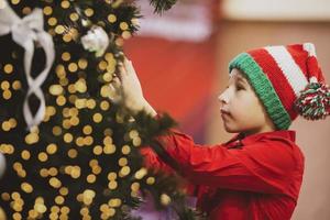 A child in a knitted Christmas hat decorates the Christmas tree. photo