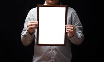 A blank diploma or a mockup certificate in the hand of a man employee wearing shirt on black background. The vertical picture frame is empty and the copy space.