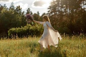 Young beautiful woman, wearing white dress, holding flowers and dancing on the meadow. Girl joying nature and freedom. Natural beauty. Dance, movement. Mental health, stress free, dreaming. Sunset. photo