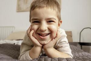 Portrait of little, smiling caucasian boy on the bed at home. Cute child relaxing, resting in bedroom. Positive emotions. Cozy and modern interior. Natural, earth colors. Casual clothes. photo