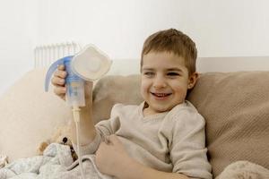 Sick little boy with inhaler for cough treatment. Unwell kid doing inhalation on his bed. Flu season. Medical procedure at home. Interior and clothes in natural earth colors. Cozy environment. photo