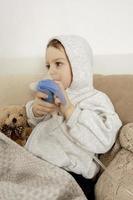 niño enfermo con inhalador para el tratamiento de la tos. niño enfermo inhalando en su cama. temporada de gripe. procedimiento médico en el hogar. interior y ropa en colores tierra naturales. ambiente acogedor. foto