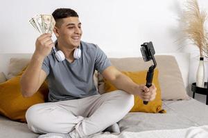 joven, blogger grabando un blog de estilo de vida, hablando con la cámara de un teléfono inteligente en un trípode y sosteniendo dinero, dólar. joven influencer filmando vlog para su canal. el hombre hace videos para seguidores en línea. foto