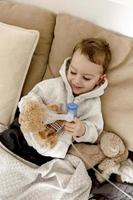Sick little boy with inhaler for cough treatment. Unwell kid doing inhalation and inhalates also his teddy bear. Flu season. Medical procedure at home. Interior and clothes in natural earth colors. photo