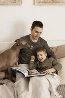 Father and son reading a book on the bed at home. Young attractive man and little boy with dog resting in bedroom. Natural earth colors. Cozy environment. Father reads a fairy tale for his child. photo