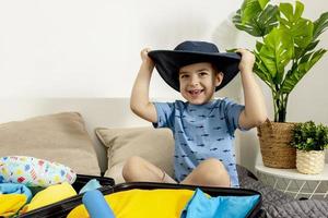 Little caucasian boy with blue shirt ready for vacation. Happy child packs clothes into a suitcase for travel. Tourist, joy of holiday. Kid at home, preparing for flying. Modern and cozy interior. photo