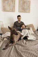 Father and son reading a book on the bed at home. Young attractive man and little boy with dog resting in bedroom. Natural earth colors. Cozy environment. Father reads a fairy tale for his child. photo