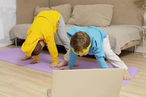 Two little caucasian kids doing fitness exercises at home. Children have fun during sport. Boys make training, looking at computer. Sport, yoga online. Workout in the apartment. Healthy lifestyle. photo