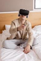 Young caucasian man sitting on bed at home with VR headset and playing interactive video game, exploring virtual reality. Man wearing VR glasses. Future, gadgets, technology, education, study online. photo