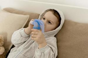 Sick little boy with inhaler for cough treatment. Unwell kid doing inhalation on his bed. Flu season. Medical procedure at home. Interior and clothes in natural earth colors. Cozy environment. photo