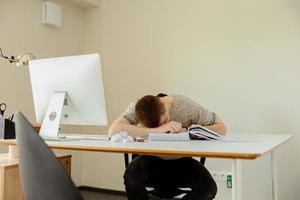 Depressed caucasian man with headache sitting at his desk in the office. Stressed young employee has problems at work. Businessman has burnout. Mental health. Overwhelmed by working. Deadline, stress. photo