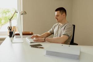 joven caucásico sentado en su escritorio en la oficina y trabajando con la computadora, respondiendo correos electrónicos. el joven empleado tiene un día de trabajo en el cargo. hombre de negocios escribiendo un plan de negocios. foto