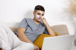 Young man with casual clothes and beauty mask on the face lying on bed with laptop computer and resting. Cosmetic for men, skin care. Man reading news in internet or watching movie. Relaxing at home. photo