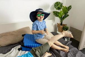 Little caucasian boy with blue shirt ready for vacation. Happy child with blue suitcase going to travel. Tourist, joy of holiday. Kid at home, preparing for flying. Modern and cozy interior. photo