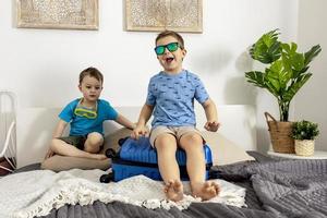 dos niños pequeños con camisas azules listos para las vacaciones. niños felices empacando ropa en una maleta para viajar. turista, alegría de vacaciones. niños en casa, preparándose para volar. interior moderno y acogedor. foto