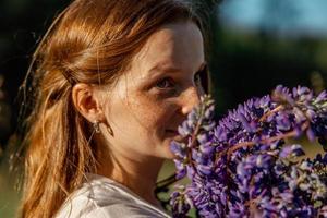 Cierra el retrato de una joven hermosa pelirroja con pecas, vestida de blanco, posando en la naturaleza. chica con cabello rojo sosteniendo flores. belleza natural. diversidad, singularidad individual. foto