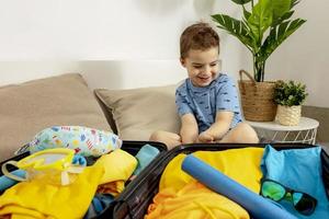 Little caucasian boy with blue shirt ready for vacation. Happy child packs clothes into a suitcase for travel. Tourist, joy of holiday. Kid at home, preparing for flying. Modern and cozy interior. photo