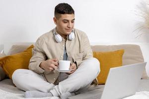 Young man with casual clothes sitting on bed with laptop computer and drinking tea or coffee. Man using notebook to surf in internet, read news, watch movie, call friends. Relaxing at home. photo