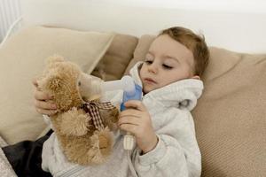 Sick little boy with inhaler for cough treatment. Unwell kid doing inhalation and inhalates also his teddy bear. Flu season. Medical procedure at home. Interior and clothes in natural earth colors. photo