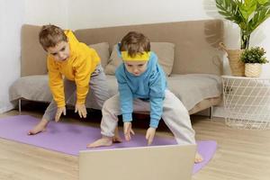 Two little caucasian kids doing fitness exercises at home. Children have fun during sport. Boys make training, looking at computer. Sport, yoga online. Workout in the apartment. Healthy lifestyle. photo
