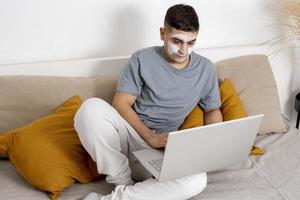 Young man with casual clothes and beauty mask on the face lying on bed with laptop computer and resting. Cosmetic for men, skin care. Man reading news in internet or watching movie. Relaxing at home. photo