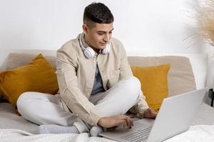 Young man with casual clothes sitting on bed with laptop computer. Man using notebook to surf in internet, play game, read news, watch movie, study or work online, call friends. Relaxing at home. photo