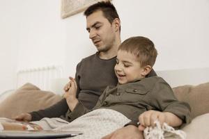 Father and son reading a book on the bed at home. Young attractive man and little boy resting in bedroom. Natural earth colors. Cozy environment. Father reads a fairy tale for his child. photo