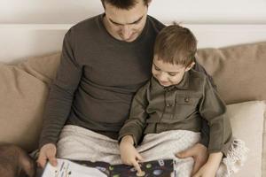 Father and son reading a book on the bed at home. Young attractive man and little boy resting in bedroom. Natural earth colors. Cozy environment. Father reads a fairy tale for his child. photo