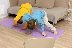 dos niños pequeños caucásicos haciendo ejercicios de fitness en casa. los niños se divierten durante el deporte. los chicos hacen entrenamiento, mirando la computadora. deporte, yoga en línea. hacer ejercicio en el apartamento. estilo de vida saludable. foto