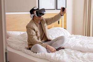 joven caucásico sentado en la cama en casa con auriculares vr y jugando videojuegos interactivos, explorando la realidad virtual. hombre con gafas vr. futuro, gadgets, tecnología, educación, estudio en línea. foto