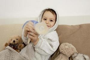 Sick little boy with inhaler for cough treatment. Unwell kid doing inhalation on his bed. Flu season. Medical procedure at home. Interior and clothes in natural earth colors. Cozy environment. photo