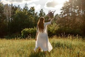 Young beautiful woman, wearing white dress, holding flowers and dancing on the meadow. Girl joying nature and freedom. Natural beauty. Dance, movement. Mental health, stress free, dreaming. Sunset. photo